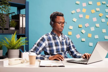 Finansal danışman bir dizüstü bilgisayar üzerinde çalışıyor, iş büyümesini ve satış yönetimini analiz ediyor. Erkek sekreter masasında otururken kulaklığı açık, cihazına bakıyor, başlangıç projesi sunumunu hazırlıyor.