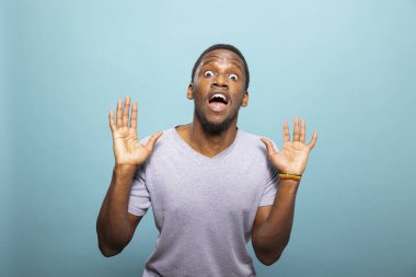 Şaşkınlık içinde iki elini de kaldırarak doğrudan kameraya bakan siyahi bir erkek. Mavi arka planda şaşırmış bir ifadeyle duran Afro-Amerikan genç adam..