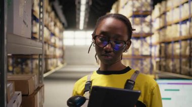 Siyah kadın personel sipariş işlemlerini sağlamak için nakliye etiketlerini tarıyor, tabletteki depo yönetim sistemindeki kayıtları güncelliyor. Depoda e-ticaret dağıtım operasyonları. Kamera A.