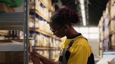 Afrika kökenli Amerikalı bir kadın ambardaki paketlere etiket yapıştırıyor, envanter ve sipariş işlemlerini yanlış etiketlerle yapıyor. Çevrimiçi büyük iş deposu alışverişi. Kamera B.