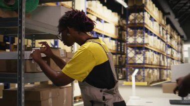 Siyahi kadın işçi paketlere yapışkan havayolu faturası etiketleri yapıştırıyor, sevkiyat detayları ve izleme bilgileriyle dağıtım için ürünleri hazırlıyor. Depoda endüstriyel operasyonlar. Kamera A.