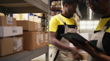 Tarayıcı kullanan siyah kadın işçi envanterdeki hava yolu faturalarını kontrol ediyor, endüstriyel depodaki lojistik operasyonları çözüyor. İşçi, raf ünitelerinin yakınındaki nakliye etiketlerini doğruladı. Kamera A.