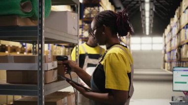 Siyah e-ticaret çalışanı takip numaralarını tarıyor, nakliye etiketlerini ve depo envanter kayıtları için ayrıntıları kaydediyor. Paletler ithalat işlemleri ile ilgileniliyor. Kamera A.