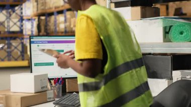 Afro-Amerikan depo memuru, takip bilgilerini bilgisayar yazılımında güncellemek için paket güzergahını denetliyor, tatbikat merkezinde yelek ve miğferle çalışıyor. Kamera B.