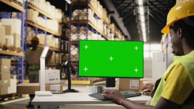 Depo ortamında çalışan siyahi bir çalışan bilgisayardaki fotokopi ekranına bakıyor. Her gün büyük raflarla çevrili endüstriyel büyük işletmeleri idare ediyor. Koruyucu donanımlı bir erkek işçi. Kamera A.