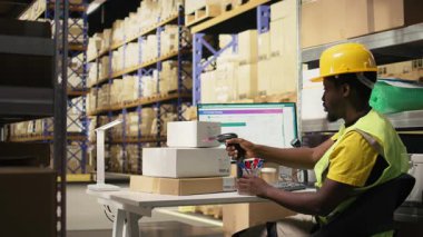 Afrikalı Amerikalı katip, büyük sanayi raflarındaki kutuların sevkiyat detaylarını kaydetmek için barkod tarayıcı kullanıyor. Paket izleme servisi için havayolu faturaları taranıyor, doğru lojistik. Kamera B.