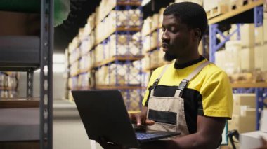 Afro-amerikan erkek personel, yoğun bakım merkezindeki depolama raflarında paketler düzenliyor. Raflarda dizüstü bilgisayarla ve ürünlerle envanter tutuyordum. E-ticaret emri işlemleri. Kamera A.