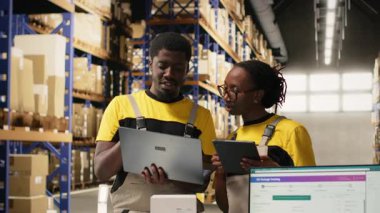 Lojistik merkezindeki sipariş işlemleri için dizüstü bilgisayardaki paket verileriyle ilgilenen siyah takım üyeleri, dağıtım protokolleri için palet taşıma etiketlerini doğruladılar. Verimli bir e-ticaret deposu. Kamera B.