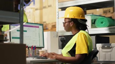 Siyah depo çalışanı dijital sevkiyat durumunu inceliyor, takip yazılımında yanlış numaralar kullanıyor. Depoda veri güdümlü sipariş takip ve teslimat güncellemeleri yapan kadın. Kamera B.