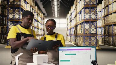 Depo personeli paketleri denetliyor. Laptop ve tabletteki bilgileri takip ediyor, sevkiyata hazırlanıyor. Rafları, karton kutuları ve işlem aletleri olan organize bir depo. Kamera A.