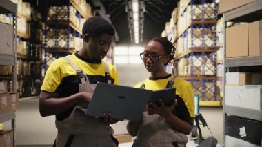 Siyahi iş arkadaşları büyük depoda sevkiyat için kutularda siparişler hazırlıyor, dizüstü bilgisayardaki envanterleri kontrol ediyor ve kargo dağıtımını yönetiyorlar. Lojistik yönetimi için paket tasnif ediyorum. Kamera A.