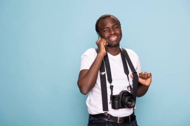 Askılı neşeli siyah adam mavi arka plan stüdyosunda dikilirken cep telefonuyla konuşuyor. Kameraya doğal ve dostane bir ifadeyle bakıyor, modern iletişimi somutlaştırıyor..