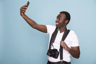 Genç siyahi adam cep telefonuyla selfie çekiyor. Boynunda bir kamera olan Afro-Amerikan erkek şahıs, cep telefonuna video kaydederken askısını tutuyor..