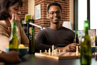 Elinde içkiyle oturan Afro-Amerikan bir adam arkadaşları arasında satranç maçı sırasında kameraya gülümsüyor. Arkadaşları masa başında bir masa oyunu oynarken mutlu siyah erkek..