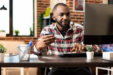 Bilgisayar başında oturan sakallı bir erkek, elinde kredi kartıyla internetten mal satın alıyor ve güvenli bir internet alışverişi yapıyor. Serbest çalışan siyahi adam evdeki banka detaylarını güncelliyor.