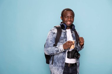 Rahat bir şekilde duran siyah bir adam, sırt çantasını tutuyor, elinde kahve fincanı, boynunda kablosuz kulaklıklar var. İzole edilmiş arka planda duran Afrikalı Amerikalı fotoğrafçının portresi.