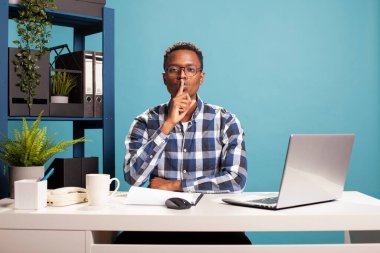 Gözlüklü genç siyahi adam, masada oturuyor ve sessiz bir jest yapıyor. Afro-Amerikalı adam işaret parmağını dudaklarına bastırıyor sessizlik işareti yapıyor, sessiz bir an istiyor, ağzı sıkı..