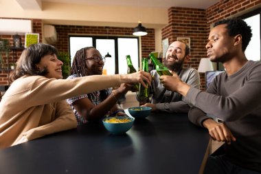 Çok kültürlü bir grup gülüyor ve bira şişelerini tokuşturuyor. Tuğla duvar odasında abur cubur ve oyunlarla rahatlıyor. Çeşitli arkadaşlar içeride eğleniyor, bira şişelerini tıkırdatıyor ve masanın etrafında atıştırıyor..