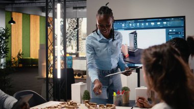 Siyahi emlak müteahhidi konut projesi yatırımını ve altyapı büyümesi stratejisini parlak bir ofiste tartışıyor, planları ve 3D modelleri analiz ediyor.