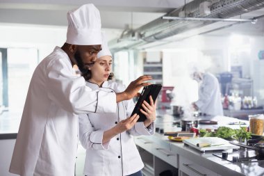 Bir tabletteki yeni gurme yemekleri için tarifleri araştıran çeşitli şeflerden oluşan bir ekip modern restoran mutfağında online bahşiş içeren yemekler hazırlıyorlar. Yemek pişirme becerisi olan uzmanlar yemek hazırlama üzerinde çalışıyorlar.