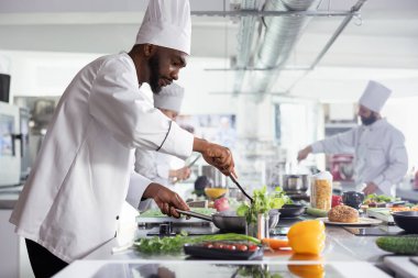 Afro-Amerikalı baş aşçı taze ürünler ve hammaddeler kullanarak paslanmaz çelik bir tavada lezzetli bir yemek pişiriyor. Modern restoran mutfağı güzel mutfak aroması ve zengin lezzetlerle dolu..