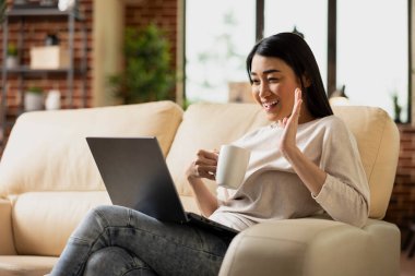 Asyalı sanal asistan elinde fincanla oturur ve dizüstü bilgisayarına el sallar, işverenle online toplantıya katılır. Gülümseyen iş kadını iş arkadaşlarını görüntülü aramada karşıladı, tartışma sırasında kahve içti