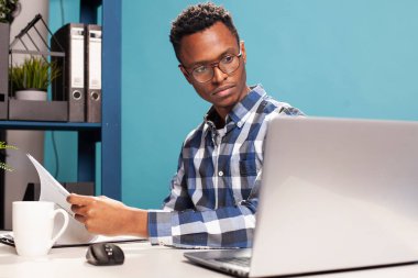 Siyahi adam, dijital cihazındaki belgelerle masasında oturuyor, okuyor ve online belgelerle karşılaştırıyor. Elinde kağıtları olan genç bir girişimci dizüstü bilgisayarına bakıyor..