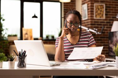 Afrika kökenli Amerikalı serbest yazar, iş müşterisiyle ciddi telefon görüşmeleri sırasında önemli belgeleri inceliyor. Elinde belgelerle odaklanmış siyah bir kadın, cep telefonuyla iş arkadaşıyla konuşuyor..