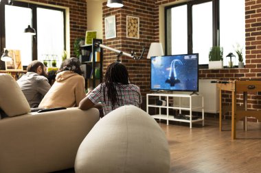 Bir grup arkadaş evde televizyon ekranının karşısında dinlenmenin keyfini çıkarıyorlar. Erkek ve kadın bireyler yumuşak mobilyaların üzerinde dinleniyor ve video oyunları oynuyorlar, kapalı alanda dostça bir rekabet içindeler..