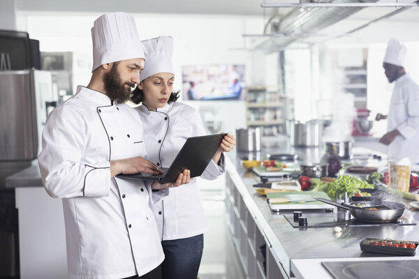 Head chefs in a white uniform researching new gourmet recipes on a laptop while overseeing food prep, chopping fresh ingredients and preparing dishes. Culinary arts in a modern kitchen.