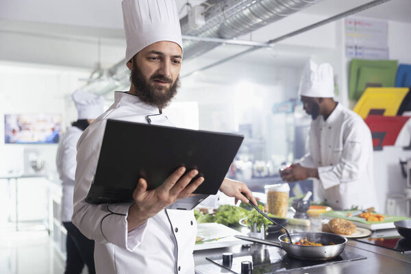 Head chef in a professional kitchen uses laptop to refine recipes, while kitchen tools and fresh ingredients fill the workspace for gourmet food prep. Culinary arts for researching dishes.