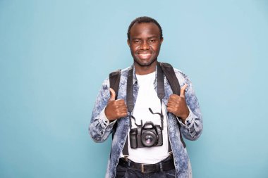 Üzerinde DSR 'ı ve sırt çantasıyla iki parmağını kaldıran neşeli siyahi adamın portresi. Kameraya bakan mutlu erkek fotoğrafçı parmaklarıyla bir anlaşma hareketi yapıyor..