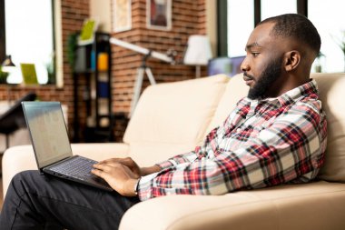 Siyahi erkek çalışan evden çalışıyor, pazarlama stratejilerini araştırıyor ve şirket raporlarını hazırlıyor. İşletme projesi sunumunu uzaktan inceleyen, dizüstü bilgisayarıyla koltukta oturan odaklanmış girişimci