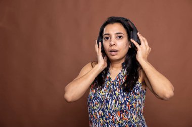 Gündelik giysiler ve kablosuz kulaklıklar takan genç bir kadın modern stüdyoda ritimle dans ediyor. Güzel Hintli kadın kameraya bakıyor ve kahverengi arka planda en sevdiği melodileri dinliyor..