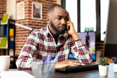 Sakallı erkek yönetici, bilgisayardaki proje güncellemelerini kontrol ederken hayal kırıklığına uğramış ve sıkılmış görünüyor. Serbest çalışan siyah adam oturma odasında dijital işlerle boğuşuyor..