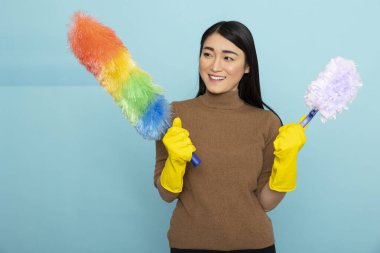 Sarı lastik eldiven giyen mutlu Asyalı kadın gökkuşağı ve beyaz toz bezi tutuyor, mavi arka planda portre için poz verirken güvenle gülümsüyor. Oda servisi ve hijyen konsepti.