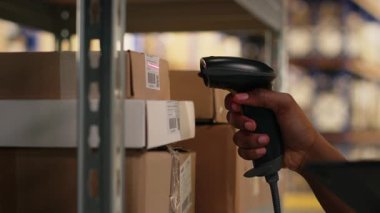 Tarayıcı kullanan siyah kadın işçi envanterdeki hava yolu faturalarını kontrol ediyor, endüstriyel depodaki lojistik operasyonları çözüyor. İşçi, raf ünitelerinin yakınındaki nakliye etiketlerini doğruladı. Kamera B.