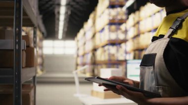 Zenci kadın tabletteki depo lojistik sistemindeki teslimat kayıtlarını kontrol ediyor. Envanter ve ambalaj faaliyetleri raf birimleri içinde gerçekleşir, doğru sıranın yerine getirilmesini sağlar. Kamera A.