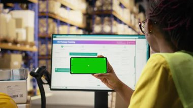 Siyah bir kadın telefonu krom anahtarla tutuyor ve izleme yazılımlarındaki paket sevkiyatını, depo rafları ve paletlerle ithalat operasyonlarını izliyor. Doğru lojistik. Kamera A.
