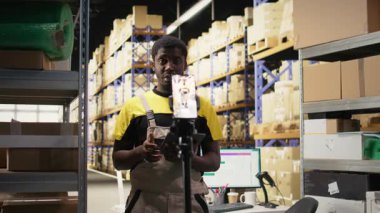 Tulumlu Afro-Amerikalı bir adam endüstriyel depoda canlı yayın sunuyor, paket taşıma ve sipariş işleme sergiliyor. Ticari bir kampanya için malları tanıtan bir işçi. Kamera A.