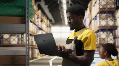 Siyah erkek personel, e-ticaret lojistik merkezindeki envanter paletlerini doğruluyor. Dizüstü bilgisayarda doğru işlem ve büyük ölçekli depo ayarlarında hızlı nakliyat için ambalaj. Kamera B.