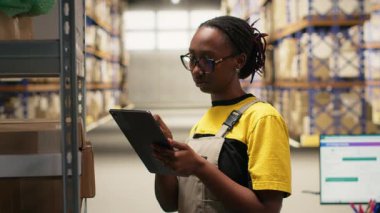 Tabletle ve dağıtım merkeziyle birlikte sipariş işleme görevlerinde çalışan siyahi kadın sanayi katibi verimli bir depolama operasyonu sağlıyor. E-ticaret dağıtımı için veri paketlerini yönetiyor. Kamera B.