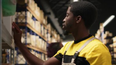 Erkek personel, sanayi deposundaki kutulara yapışkan nakliye etiketleri yapıştırıyor, paketleri yanlış takip numaralarıyla etiketliyor ve sorunsuz ekspres teslimat ve sipariş işlemlerini sağlama alıyor. Kamera B.