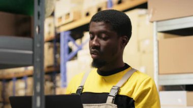 Afro-amerikan erkek personel, yoğun bakım merkezindeki depolama raflarında paketler düzenliyor. Raflarda dizüstü bilgisayarla ve ürünlerle envanter tutuyordum. E-ticaret emri işlemleri. Kamera B.