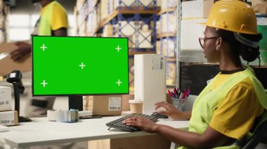 Güvenlik yeleği giymiş bir kadın bilgisayardaki yeşil ekranın yanındaki barkodları kontrol ediyor, e-ticaret dağıtım operasyonları üzerinde çalışıyor ve kask takıyor. Büyük bir depo rafı. Kamera B.