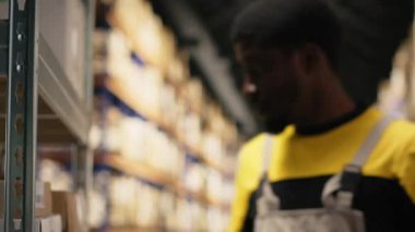 Siyah erkek işçi, envanterdeki hava yolu faturalarını kontrol ederek endüstriyel depodaki lojistik operasyonları çözüyor. İşçi, raf ünitelerinin yakınındaki nakliye etiketlerini doğruladı. Kamera B.