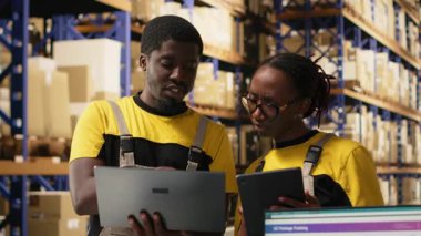 Depo personeli paketleri denetliyor. Laptop ve tabletteki bilgileri takip ediyor, sevkiyata hazırlanıyor. Rafları, karton kutuları ve işlem aletleri olan organize bir depo. Kamera B.