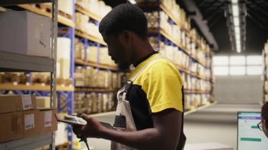 Siyah erkek işçi, envanterdeki hava yolu faturalarını kontrol ederek endüstriyel depodaki lojistik operasyonları çözüyor. İşçi, raf ünitelerinin yakınındaki nakliye etiketlerini doğruladı. Kamera A.