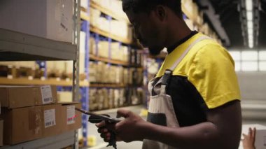 Envanterdeki barkod etiketlerini kaydetmek için tarayıcı kullanan Afrikalı Amerikalı bir adam endüstriyel depodaki lojistik operasyonları çözüyor. İşçi, depo raflarının yakınındaki nakliye etiketlerini kontrol ediyor. Kamera A.