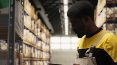 Envanterdeki barkod etiketlerini kaydetmek için tarayıcı kullanan Afrikalı Amerikalı bir adam endüstriyel depodaki lojistik operasyonları çözüyor. İşçi, depo raflarının yakınındaki nakliye etiketlerini kontrol ediyor. Kamera B.